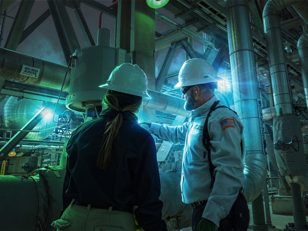 Two workers in white hard hats, seen from behind, reviewing insulated piping and equipment inside a power generation plant