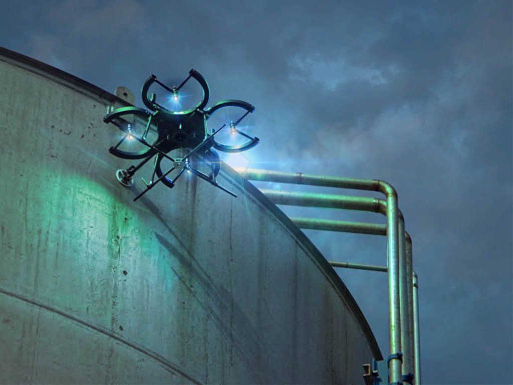 Caged inspection drone with lights hovering against the curved wall of a storage tank at dusk
