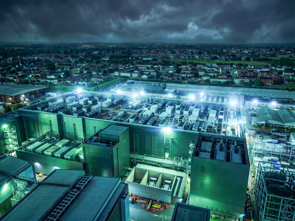 Aerial view at dusk of a large floodlit industrial facility with rooftop equipment and steel structures, a town beyond