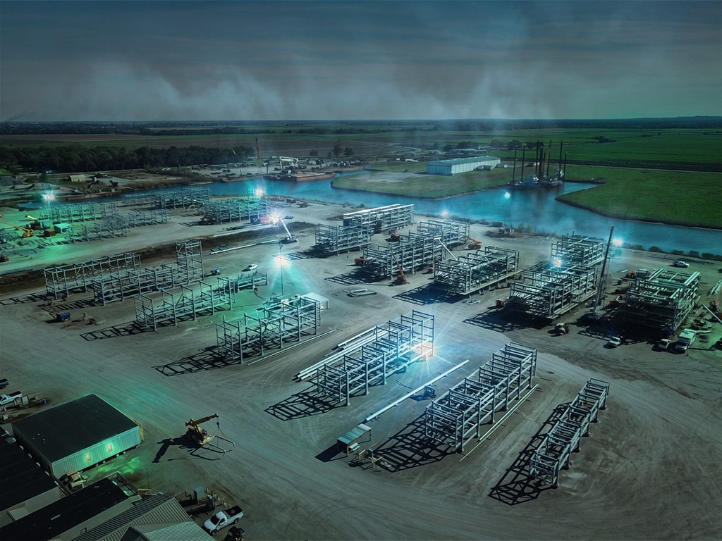 Aerial view at dusk of a floodlit fabrication yard with rows of structural steel modules beside a waterway