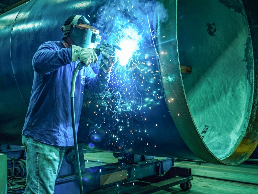 Welder in a blue work shirt welding a large steel vessel shell, sparks spraying across its surface