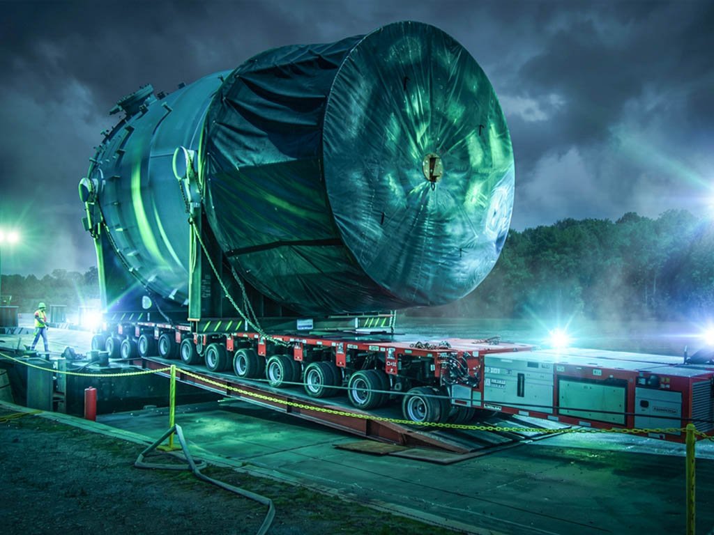 Massive wrapped pressure vessel loaded on a multi-axle self-propelled transporter at dusk, floodlit under a stormy sky