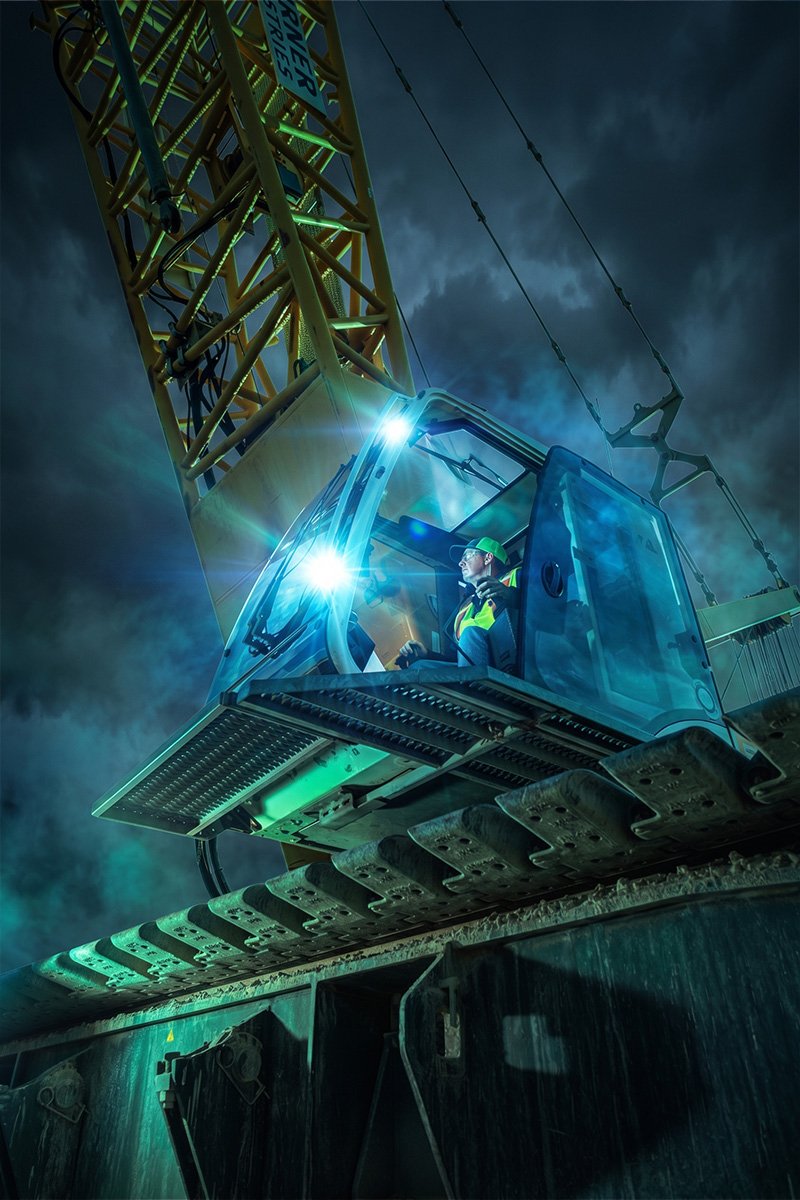 Crane operator in a green hard hat at the controls of a crawler crane cab, lit against a stormy night sky